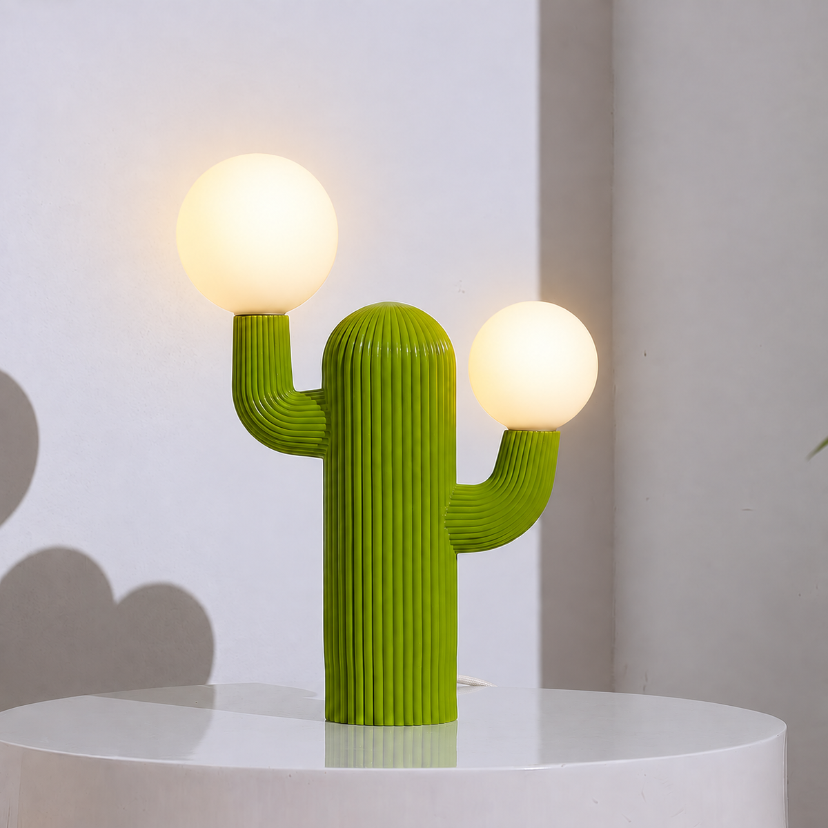 A cactus-shaped table lamp with two glowing spherical bulbs, shown on a white pedestal against a light gray wall.