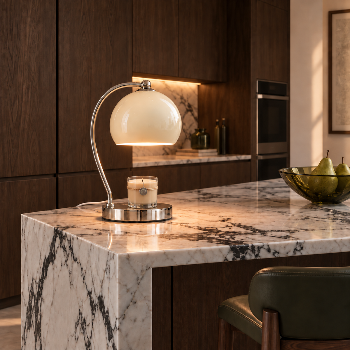 Cream Wick table lamp illuminating an unlit glass candle with twine and a silver seal on a white-and-black marble kitchen island, with dark oak cabinetry and green accents.