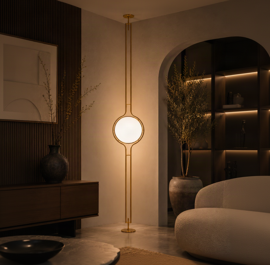 A modern floor lamp with a glowing circular shade and slim gold-toned metal frame, standing in a warmly lit contemporary living room.