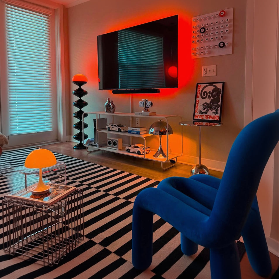 A modern living room corner featuring a black and white striped rug, a bright blue armchair in the foreground, a glass media console with a wall-mounted TV, and dramatic red and orange ambient lighting on a beige wall.