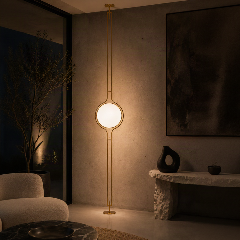 A modern floor lamp with a long gold metal frame and a glowing oval light bulb, placed in a warmly lit contemporary living room corner.
