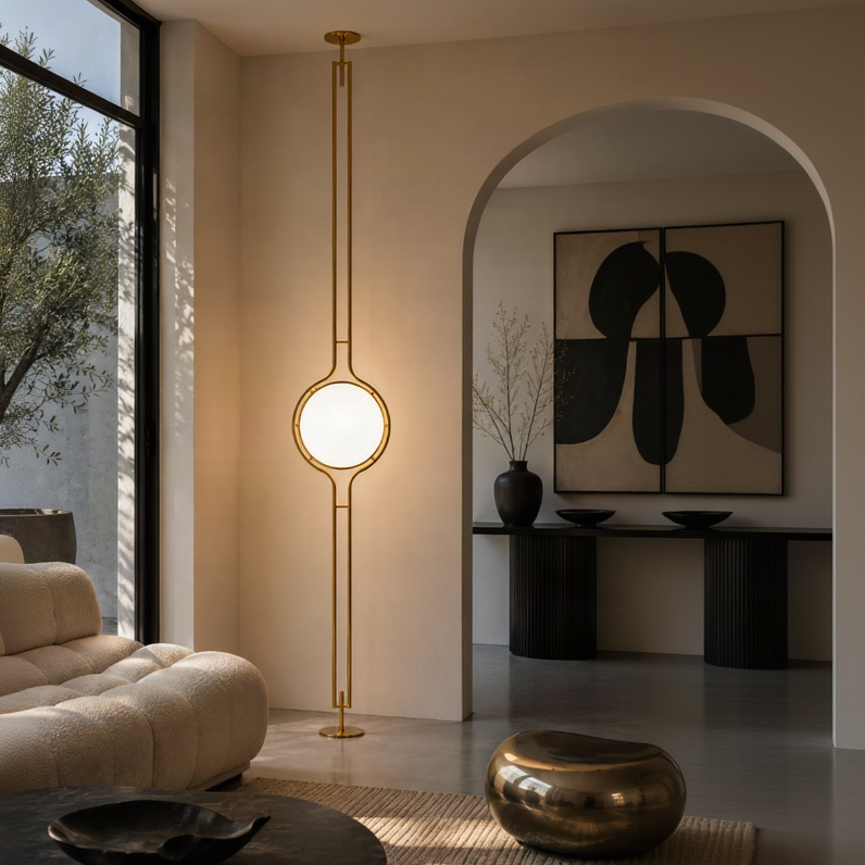 A modern brass floor lamp with a circular glowing light in the center, suspended by a slim vertical rod in a warm, contemporary living room.