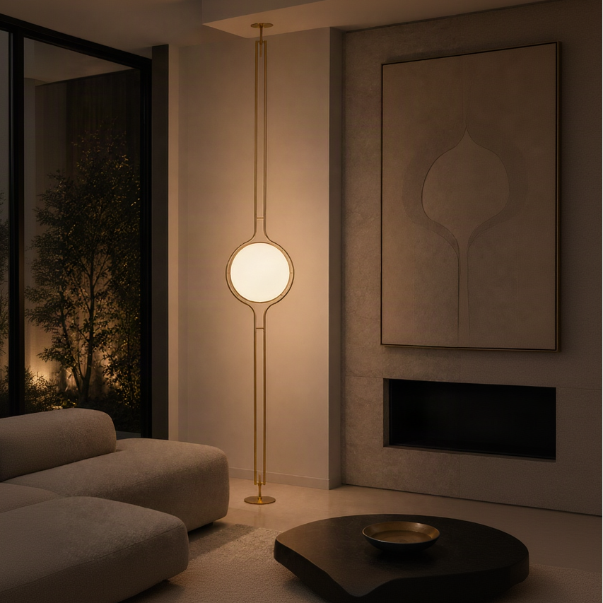 A modern floor lamp with a tall brass frame and a glowing oval shade, positioned in a warm, contemporary living room next to a gray sofa and textured wall.