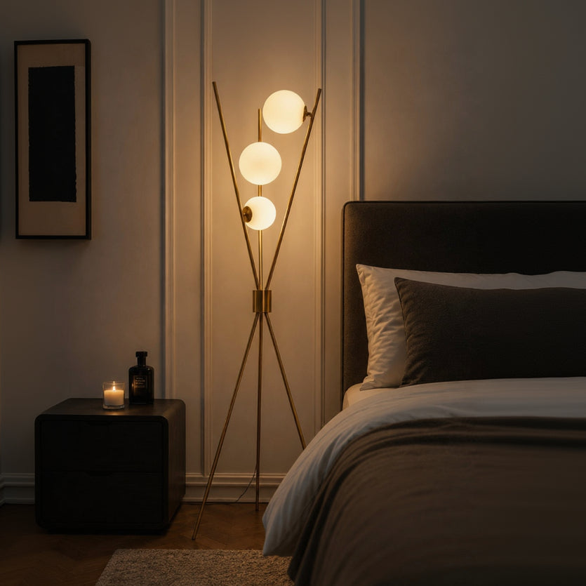 A modern floor lamp with three glowing spherical bulbs on slim angled metal legs, placed beside a bed in a dimly lit bedroom.