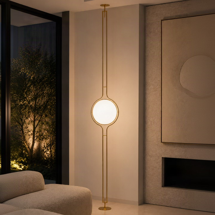 A modern floor lamp with a slender gold metal frame and a glowing oval light bulb, positioned in a contemporary living room next to a beige sofa and a dark coffee table.