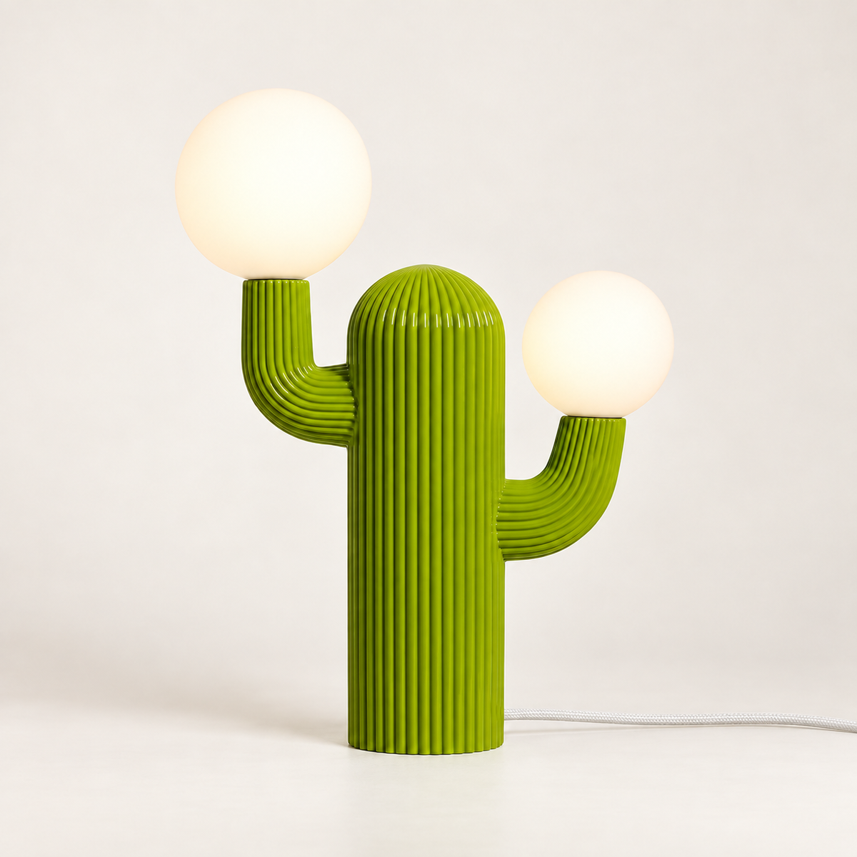Prickly green cactus table lamp illuminated in warm white with a white woven fabric cord on an ivory studio background.